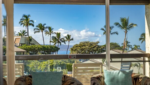 A balcony with a view of palm trees and a building across the water. - Cozy Beachside Retreat Steps from Kamaole III (Kihei)