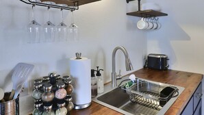 Una cocina con encimera de madera, pileta de acero inoxidable y un tostador. Hay estantes con frascos y tazas de vidrio, y un portavasos con copas de vino.