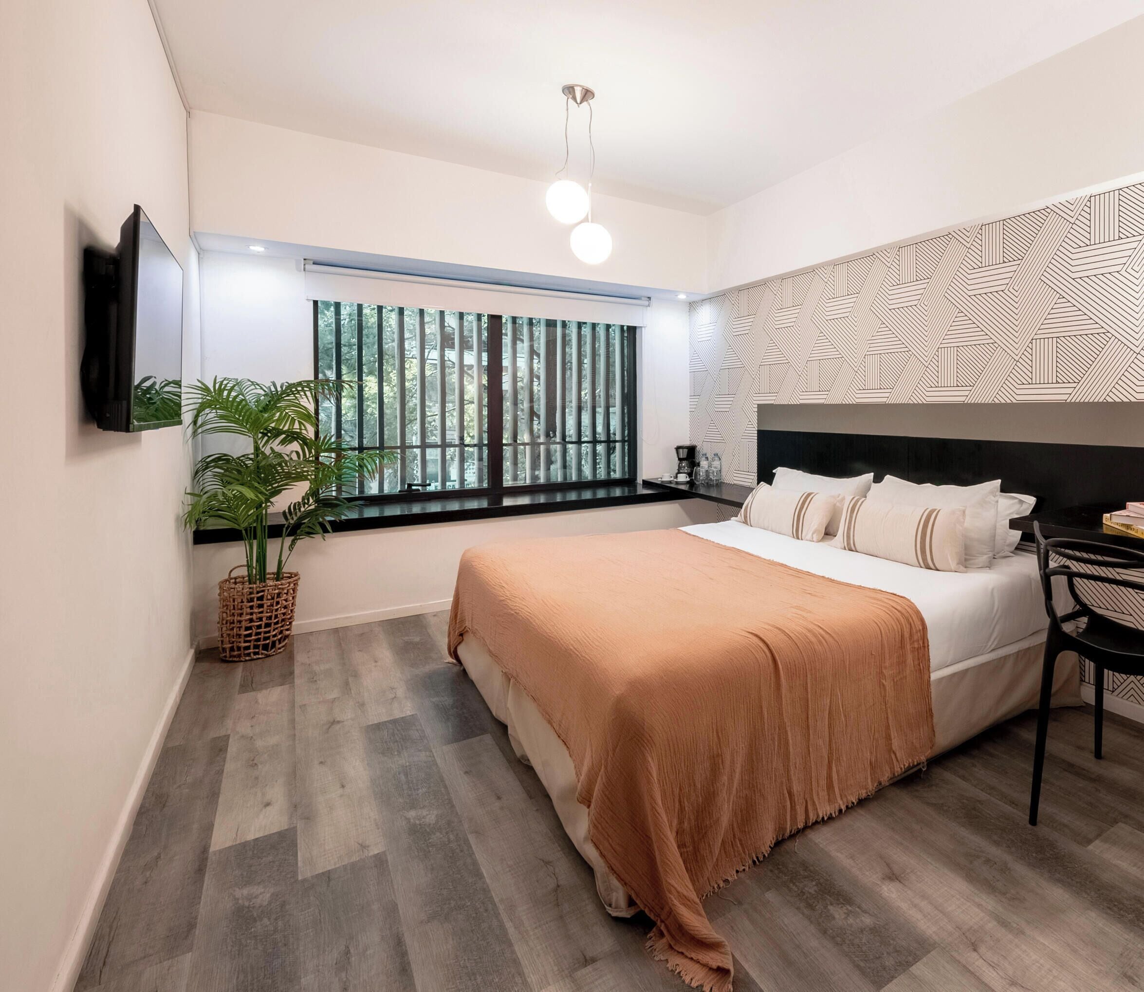 Un dormitorio moderno con cama, televisor montado en la pared, ventana con persianas, pared con estampado y una lámpara en el techo.
