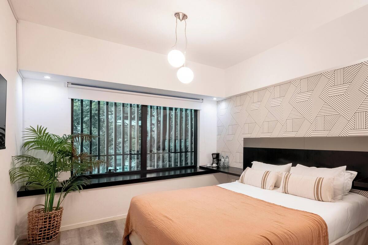 A modern bedroom with a large bed, a window with a view of trees, a geometric patterned wall, and a hanging light fixture.