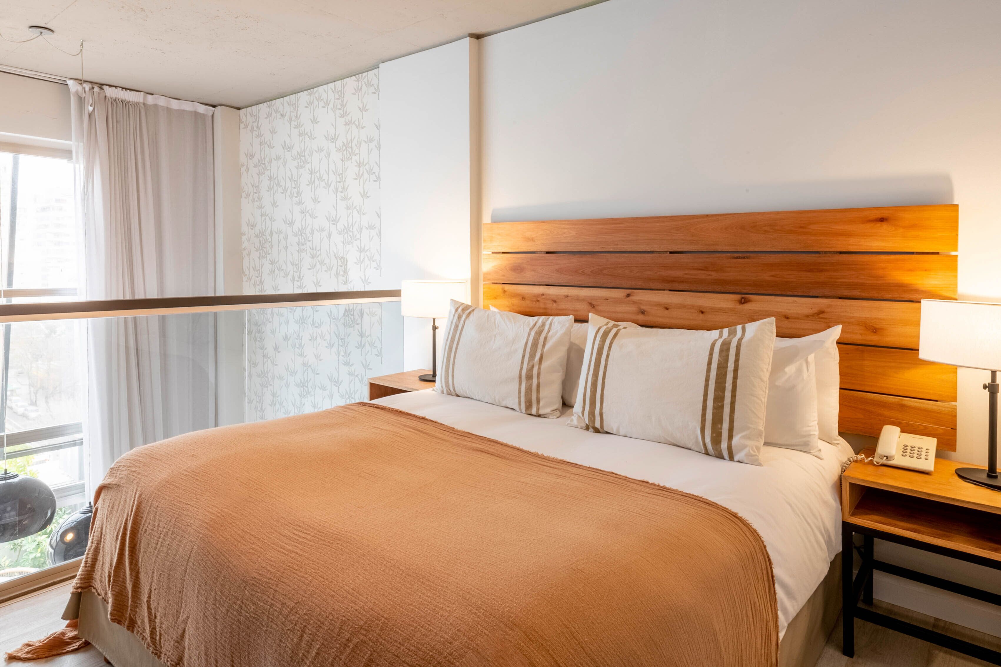 A neatly made bed with a wooden headboard, white bedding, and a brown throw blanket. There is a bedside lamp and a phone on a wooden nightstand.