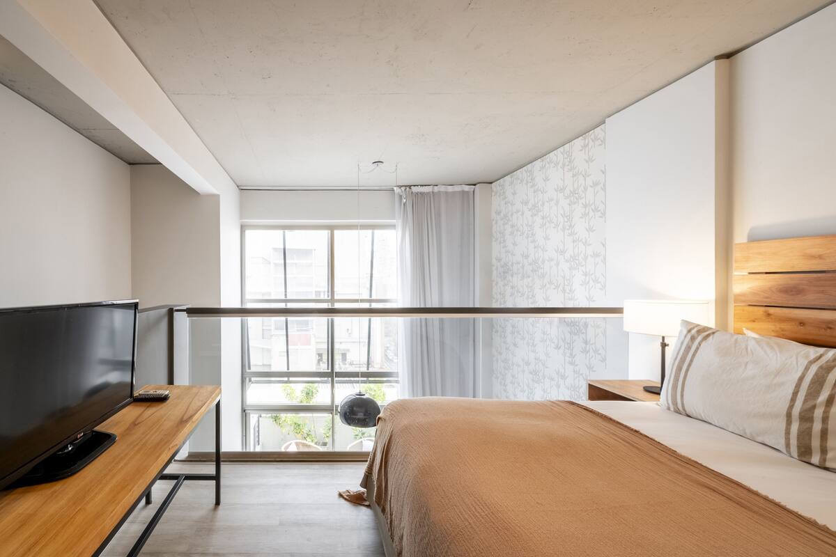 A bedroom with a bed, a TV, a window with curtains, and a view of the outside.