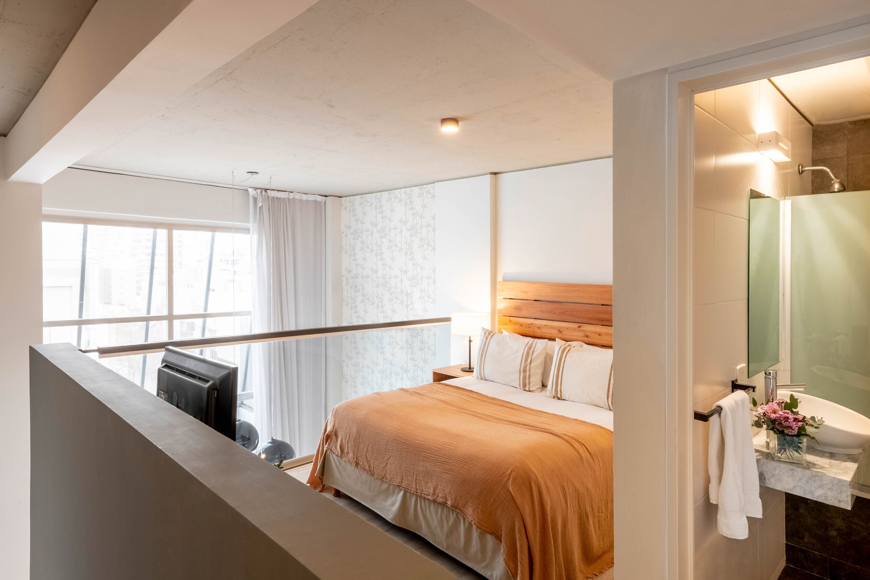 A hotel room with a large bed, a bathroom with a mirror and sink, and a balcony with a chair.