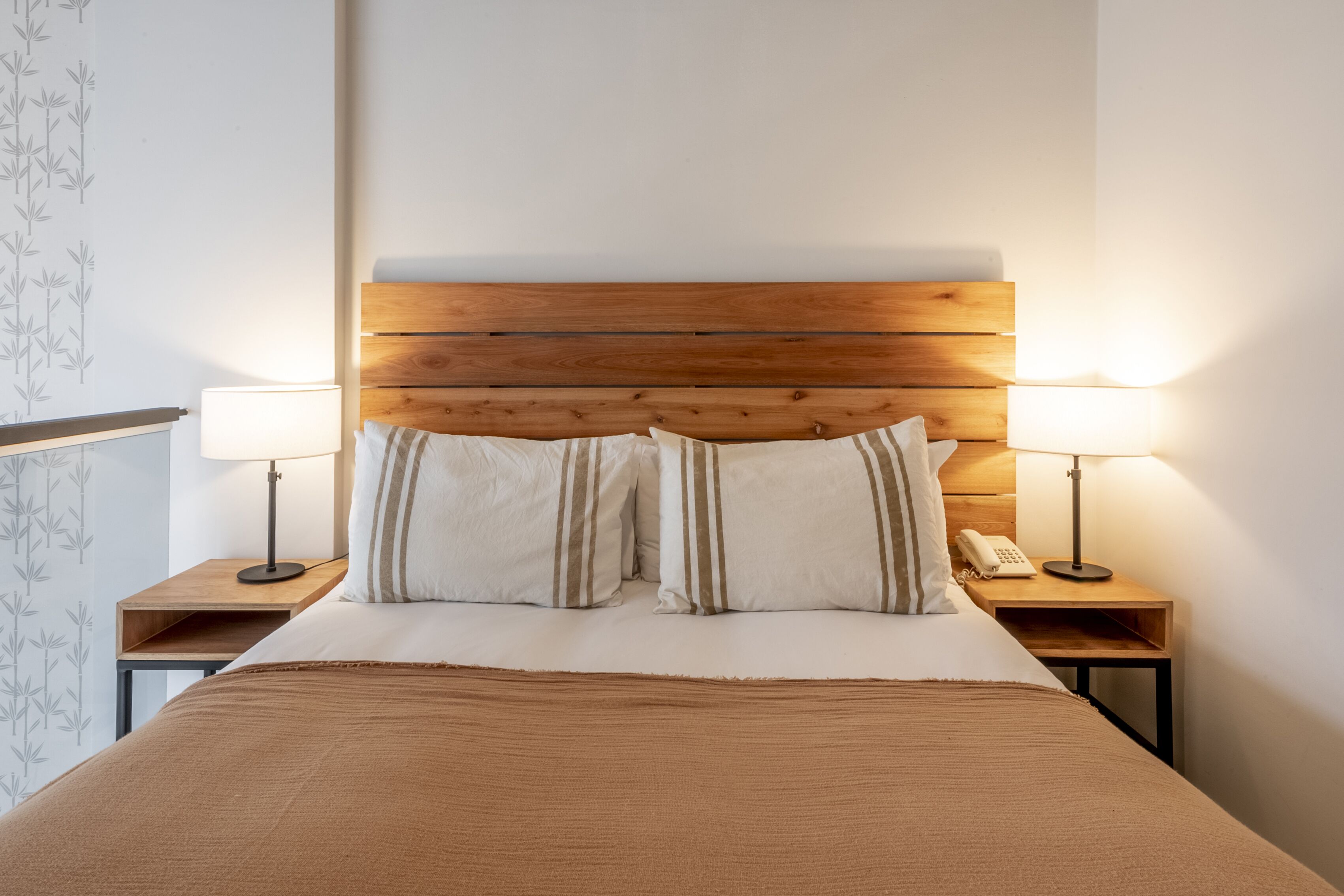 Una cama bien hecha con cabecera de madera, dos mesitas de noche con lámparas y un teléfono en una de ellas.