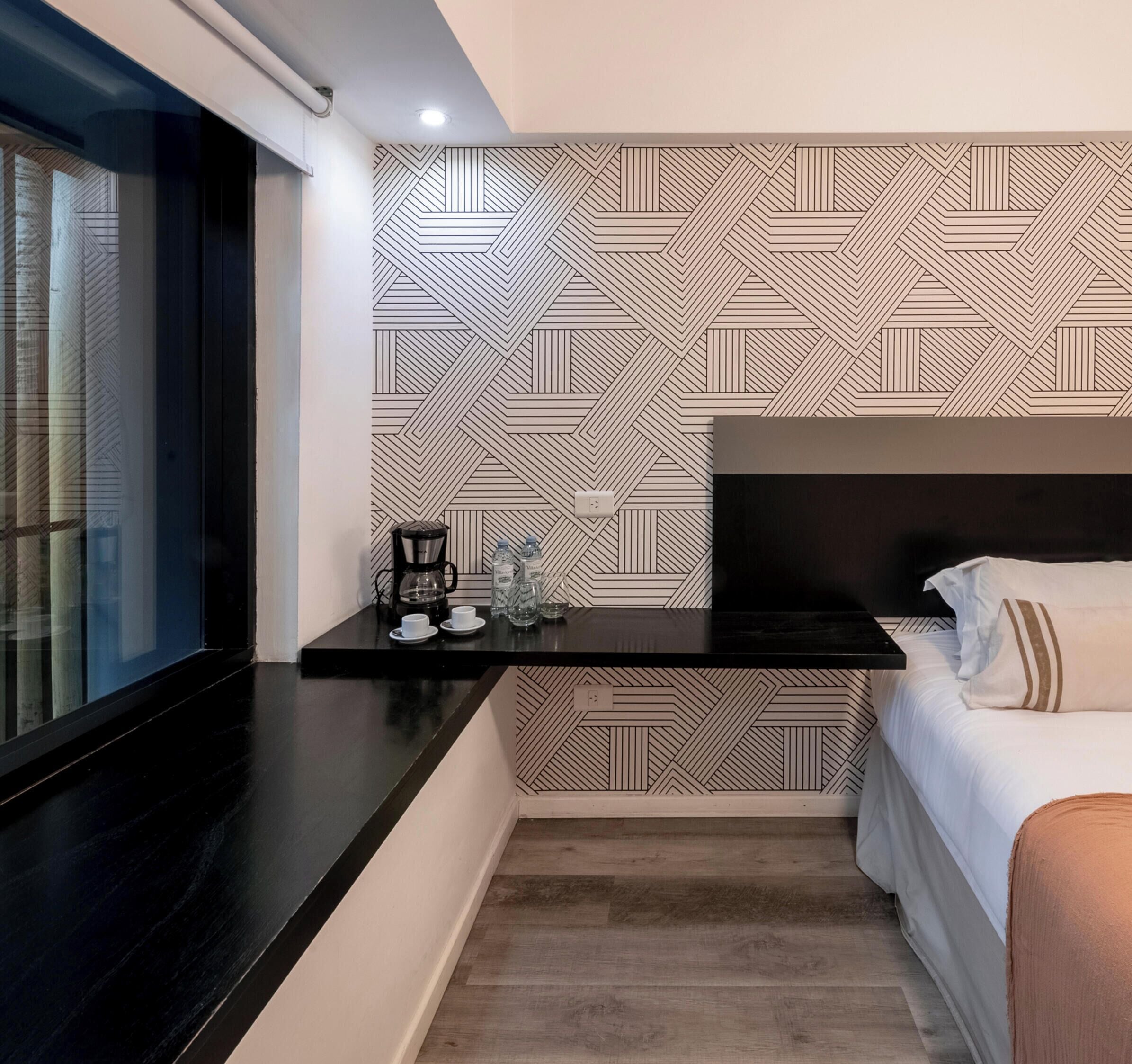 A modern hotel room with a black countertop, a bed with white bedding, a patterned wall, and a window with a view.