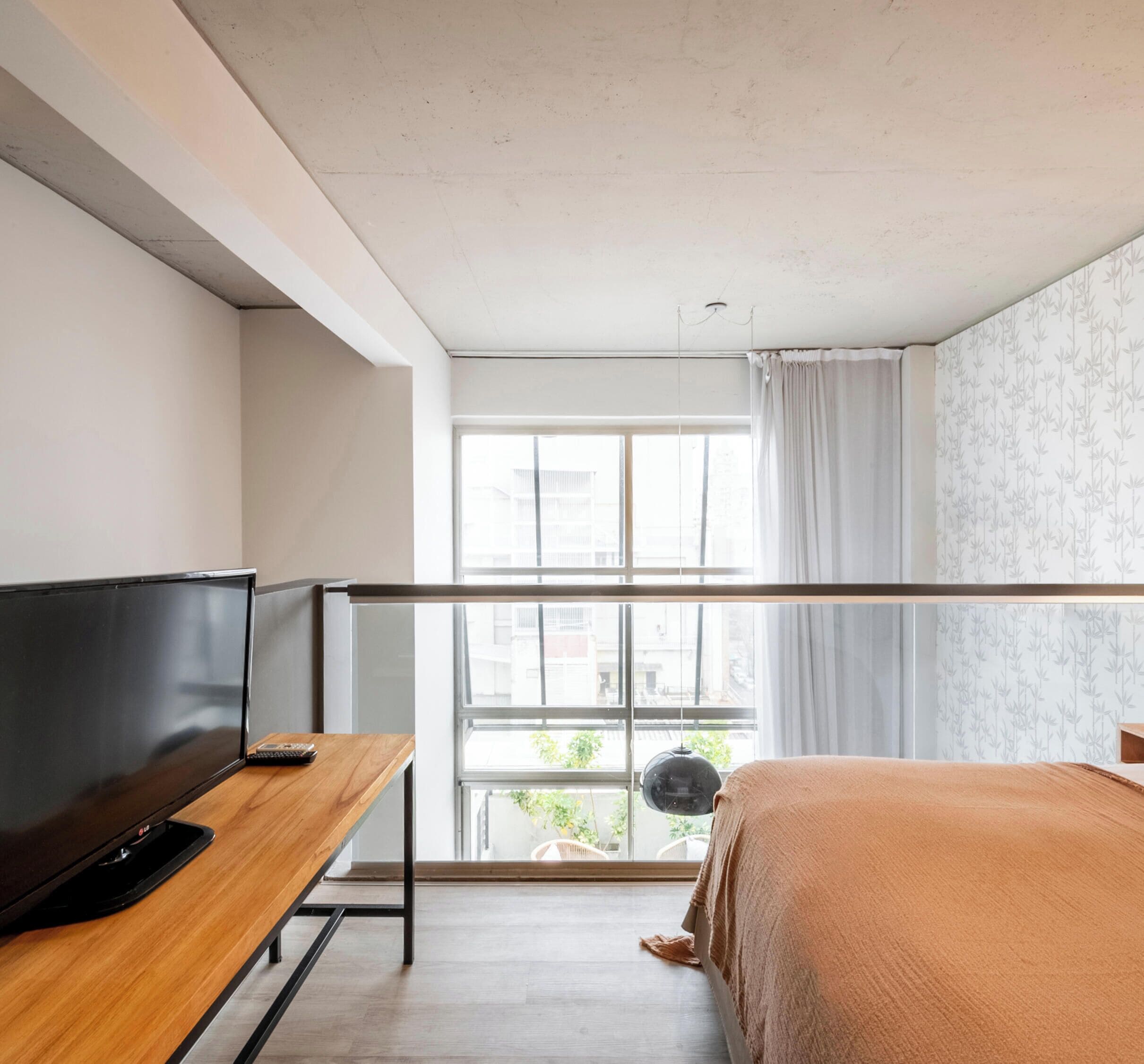 Un dormitorio con cama, televisión sobre una mesa de madera, ventana con cortinas y vistas a edificios.