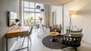 Un salon moderne avec une table basse en bois, un canapé, un téléviseur fixé au mur et une grande fenêtre donnant sur l’extérieur.