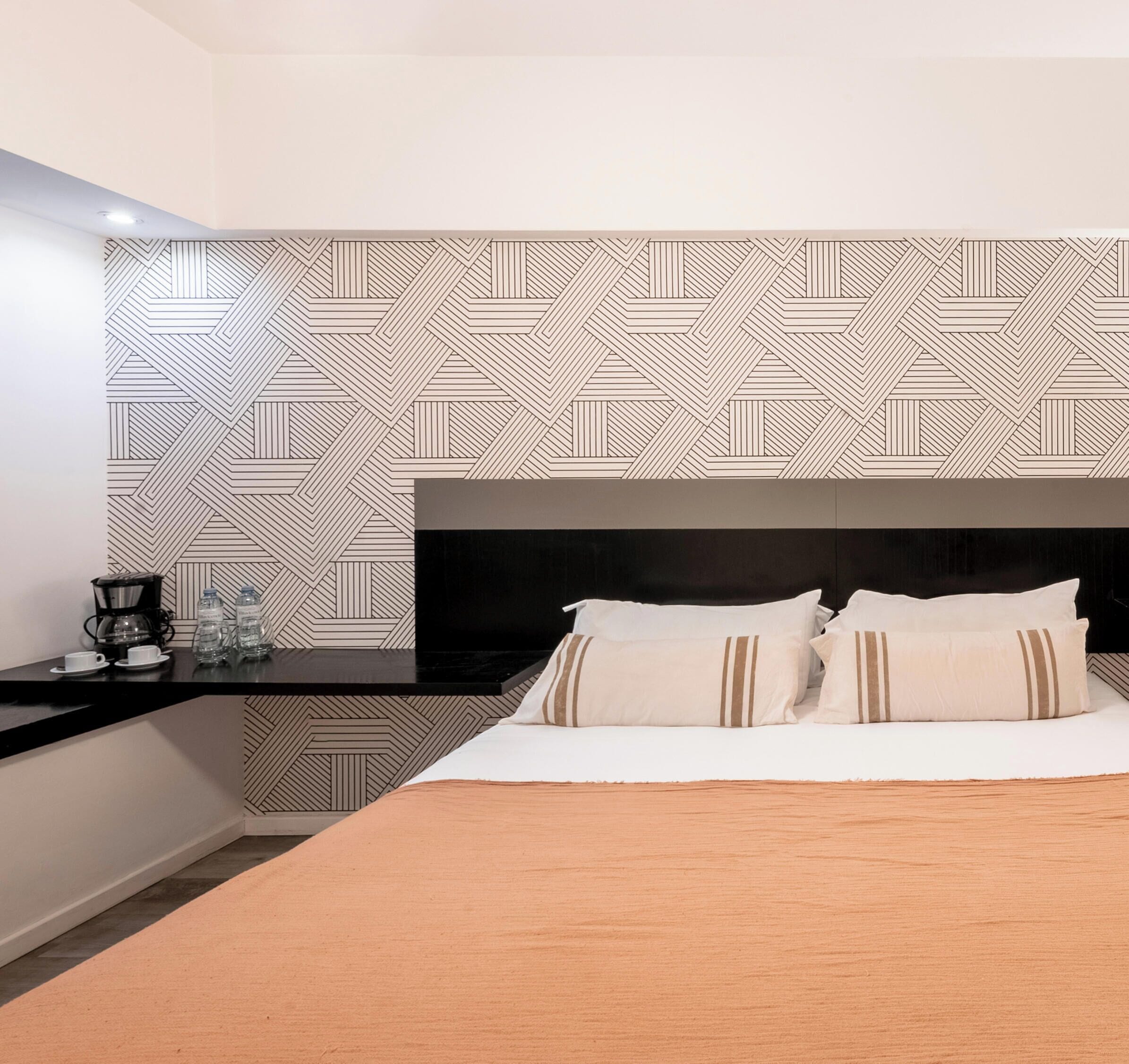 A modern hotel room with a geometric patterned wall, a black headboard, a bed with white and brown striped pillows, a wooden bedside table with a coffee maker and water bottles, and a dark floor.