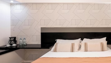 A modern hotel room with a geometric patterned wall, a black headboard, a bed with white and brown striped pillows, a wooden bedside table with a coffee maker and water bottles, and a dark floor.