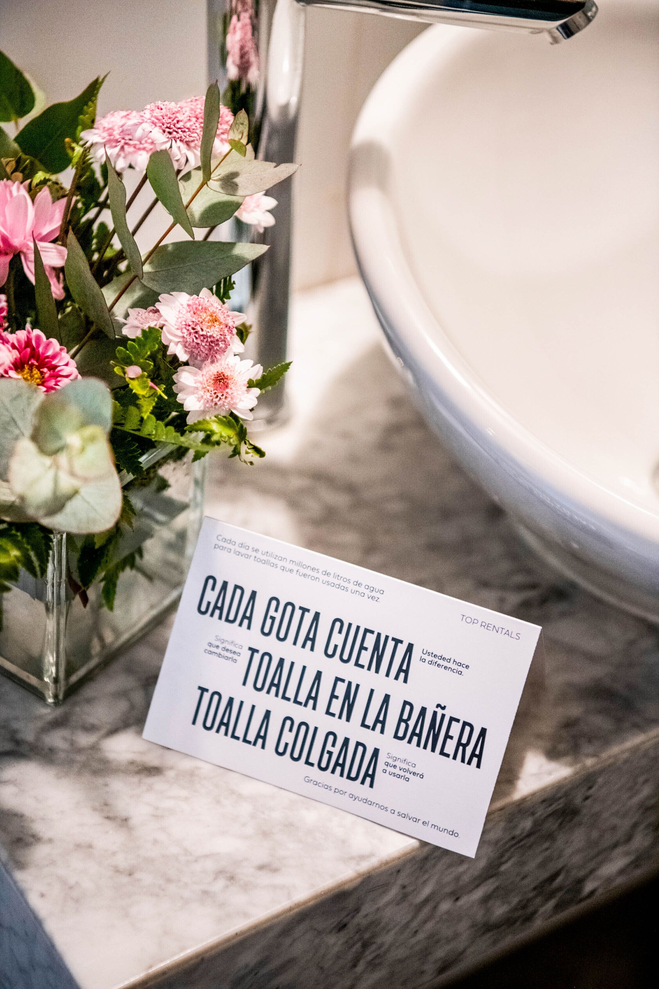 A bathroom with a marble countertop, a vase of pink flowers, and a sign in Spanish.