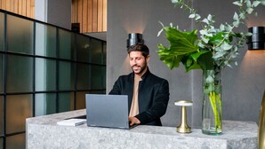 Un comptoir d’accueil avec un ordinateur portable, un vase contenant des feuilles vertes et un intérieur moderne agrémenté de panneaux en bois et d’une cloison en verre.