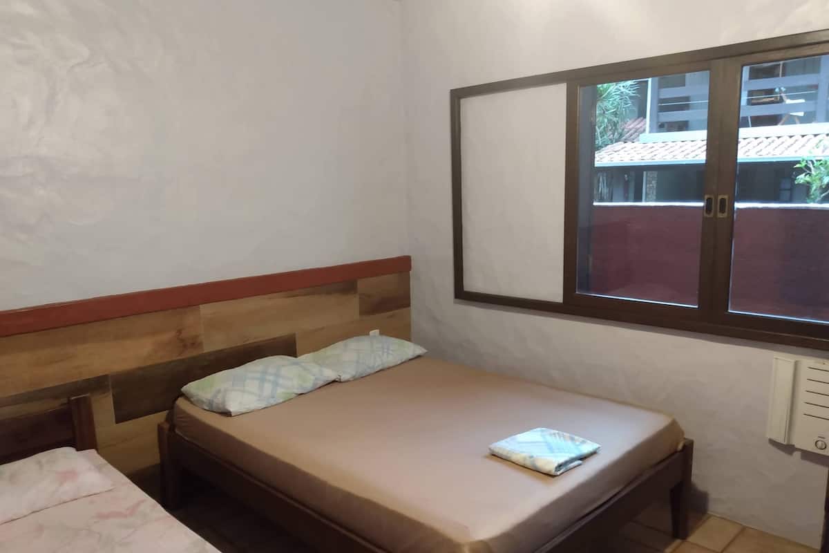 Una cama individual con cabecera de madera, una ventana con vistas a edificios y un enchufe fijado a la pared.