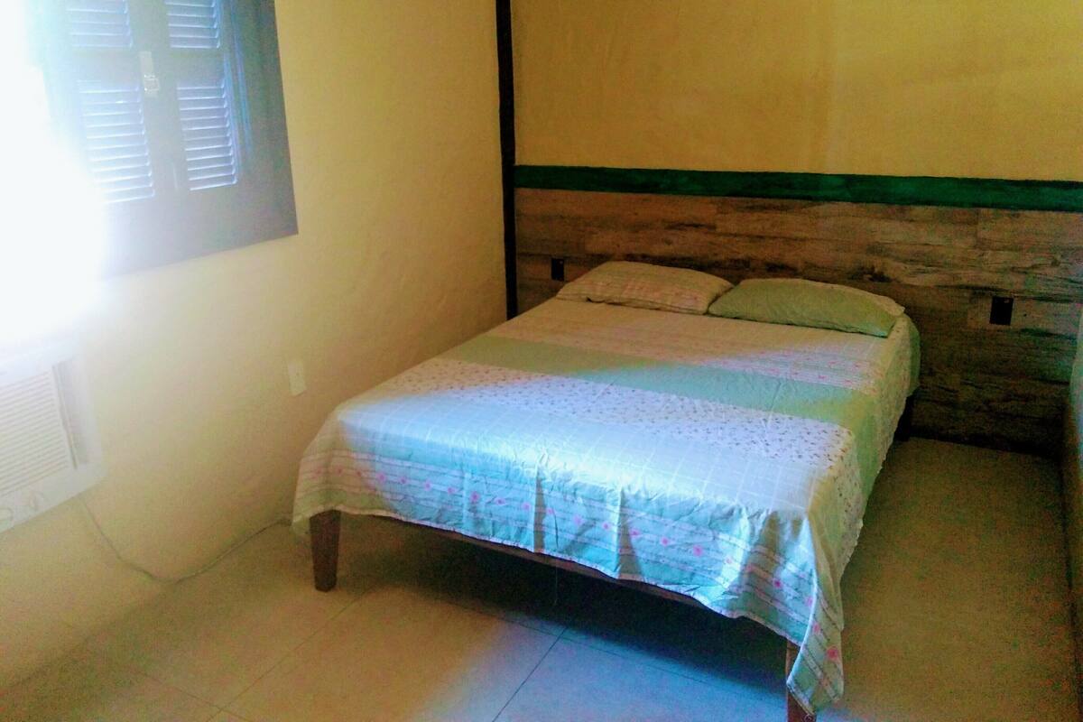 Una habitación pequeña con una cama individual, cabecera de madera, ventana con persianas y pared sencilla.