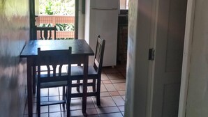 A small kitchen with a refrigerator, a dining table with chairs, and a view of a garden through a window. - Condomínio Sargento Laranja (Garopaba)