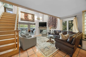 A spacious living area with a staircase, a fireplace, a television, and a large window with curtains.