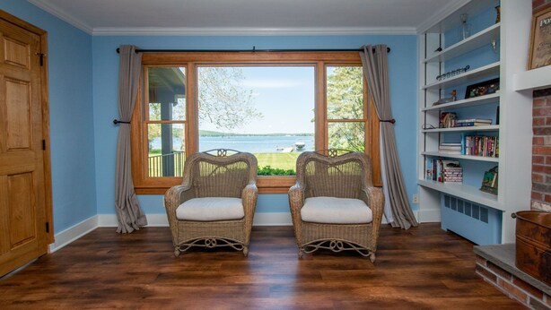 A room with two wicker chairs facing a large window overlooking a lake, a bookshelf to the right, and wooden flooring. - Maya's Palace: Pet-Friendly Keuka Lake Escape! (Keuka Park)