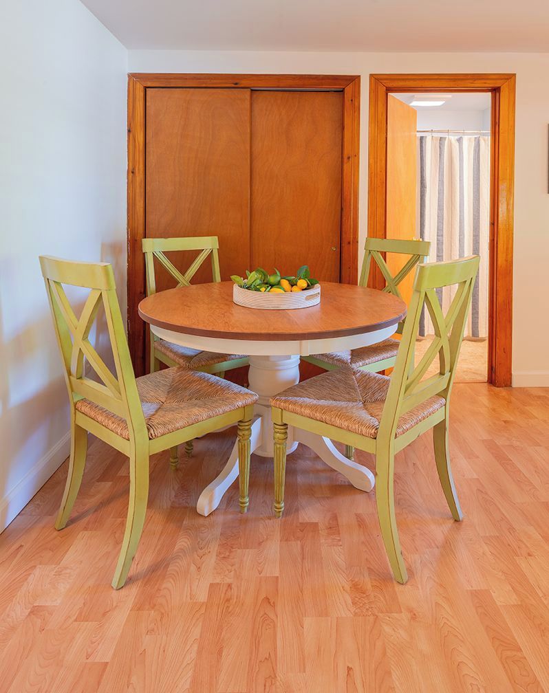 A dining area with a round table and four chairs, a wooden door in the background, and a view of a bathroom through an open door.