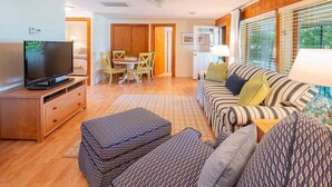 Ein gemütliches Wohnzimmer mit Fernseher, einer Couch, einem Esstisch mit Stühlen und einem Fenster mit Jalousien.