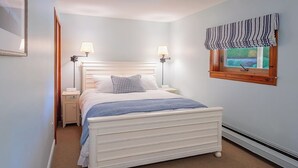 Ein Schlafzimmer mit einem weißen Bett, zwei Nachttischlampen, einem Fenster mit einem gestreiften Vorhang und einem kleinen Nachttisch.