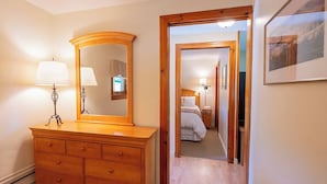 Een houten dressoir met spiegel en lamp, een gang die leidt naar een slaapkamer met een bed en nachtkastje, en een lijst met foto aan de muur.