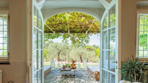An open patio with a stone floor, white furniture, and a view of trees and a pergola.
