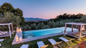 A private pool area with lounge chairs, a pergola, and a view of the city at dusk.