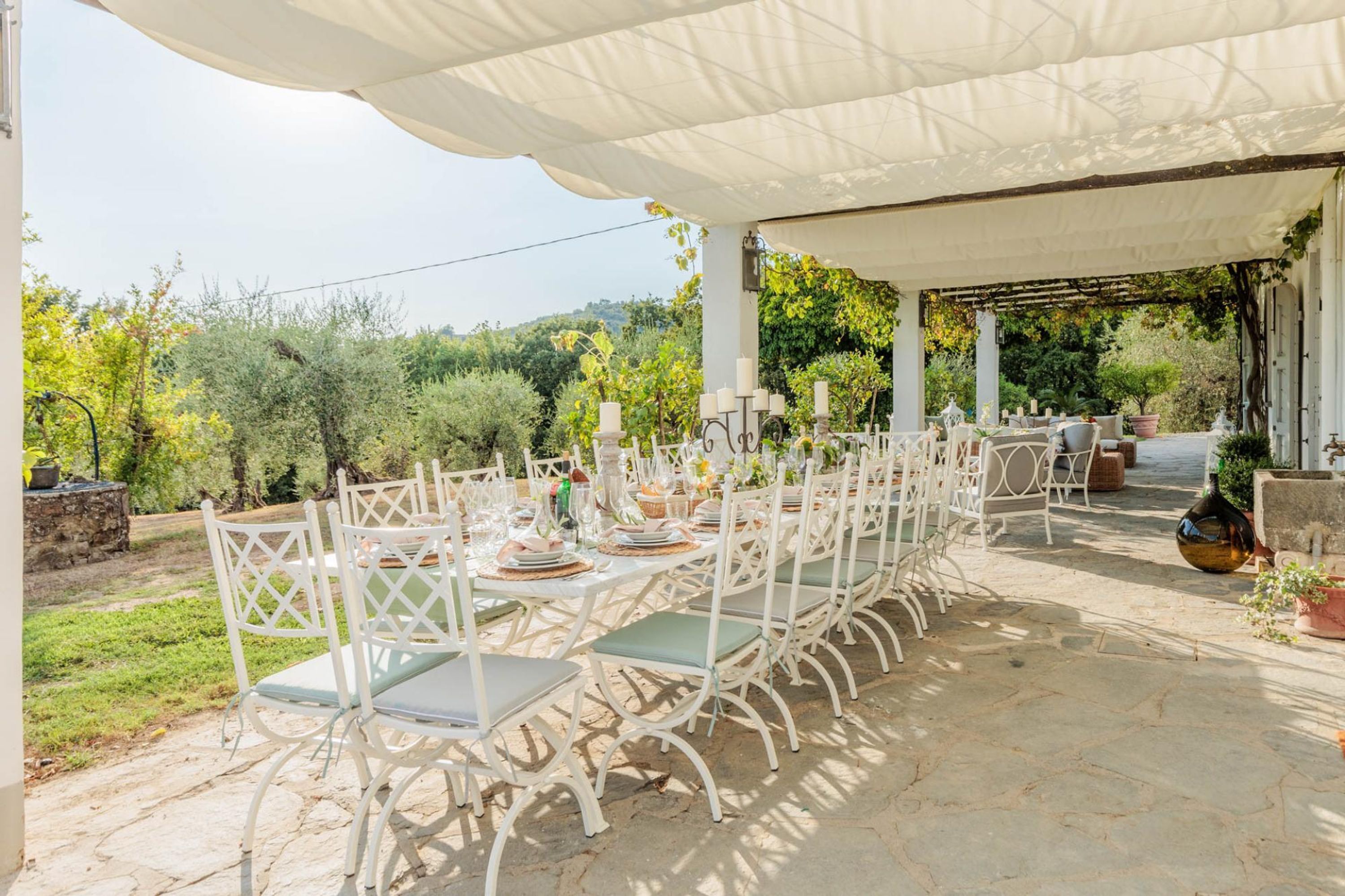 An outdoor dining area with white metal chairs and tables, covered by a white canopy, set against a backdrop of greenery and trees.