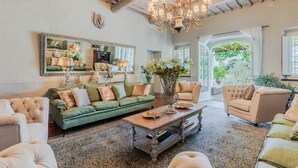 A spacious living room with a green sofa, beige armchairs, and a wooden coffee table. A large mirror and chandelier are visible, along with a view of the outdoors through large windows.