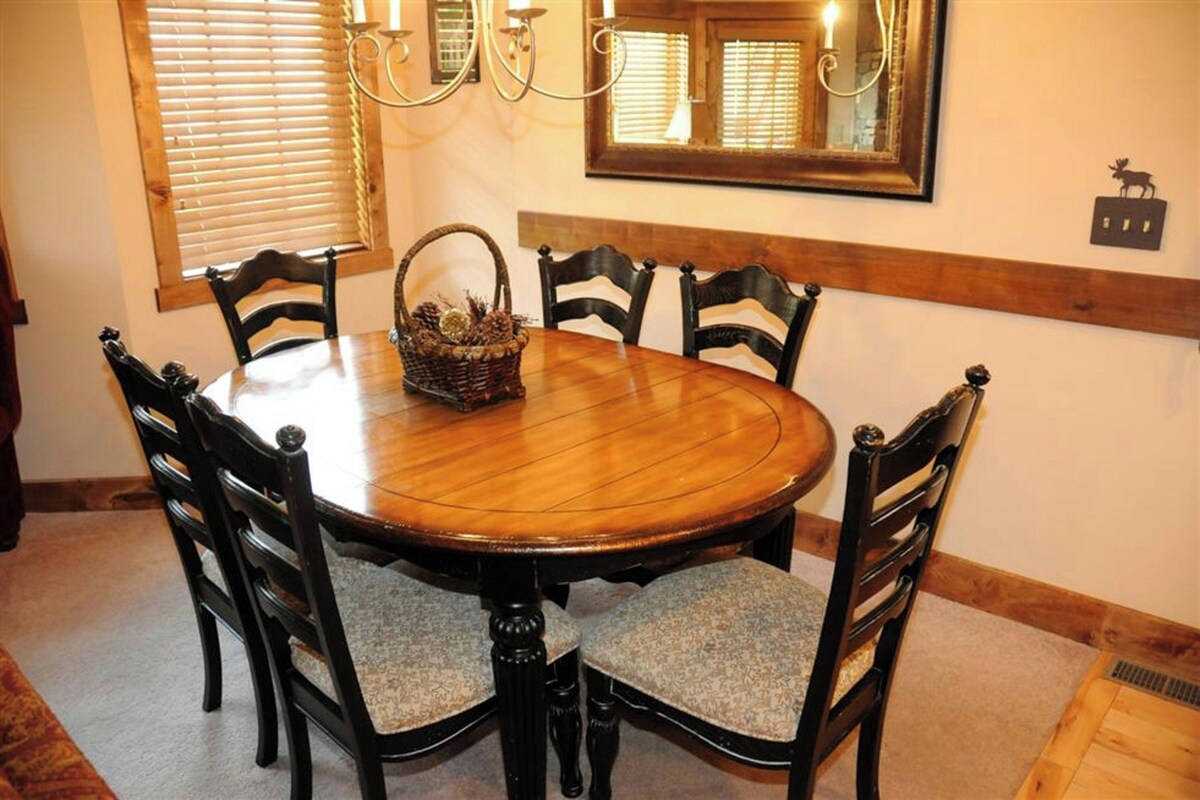 A dining room with a wooden table surrounded by six chairs, a woven basket on the table, and a chandelier above.