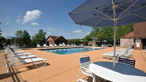 Un espace piscine avec des chaises longues et des parasols, un petit bâtiment et une zone délimitée par une clôture, plantée d’arbres.