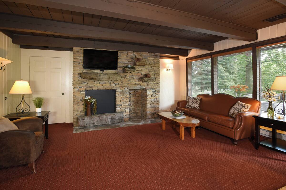 A cozy living room with a stone fireplace, leather sofa, and a large window with blinds.