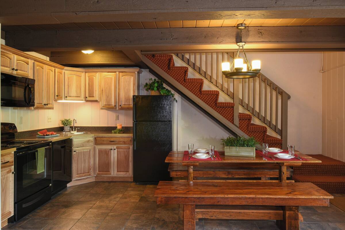 A kitchen with wooden cabinets, a black refrigerator, a dining table set for four, and a staircase with a red patterned carpet.
