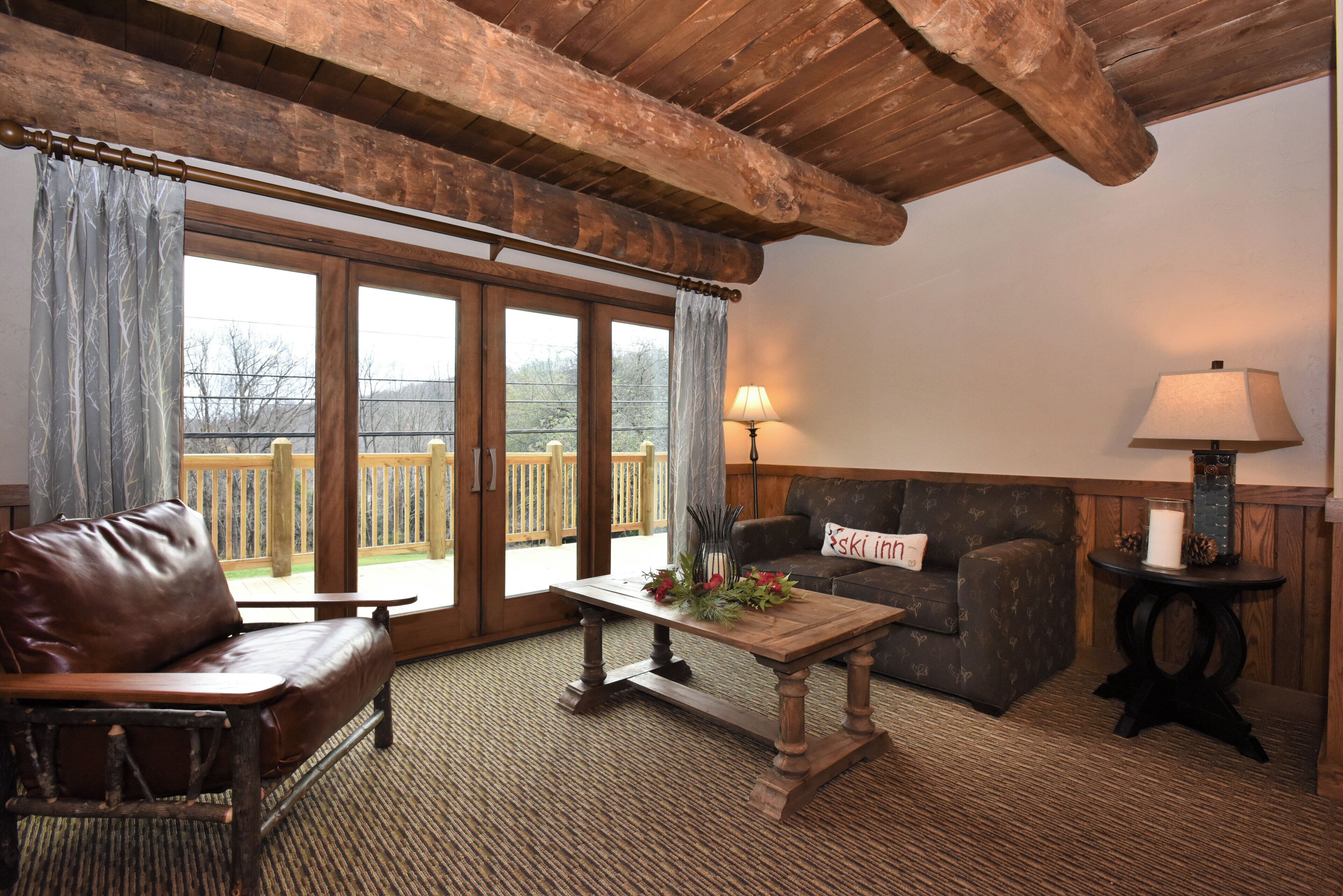 A cozy living room with a wooden ceiling, a leather sofa, a wooden coffee table with a floral arrangement, a stone fireplace, and a view of a wooden deck through glass doors.