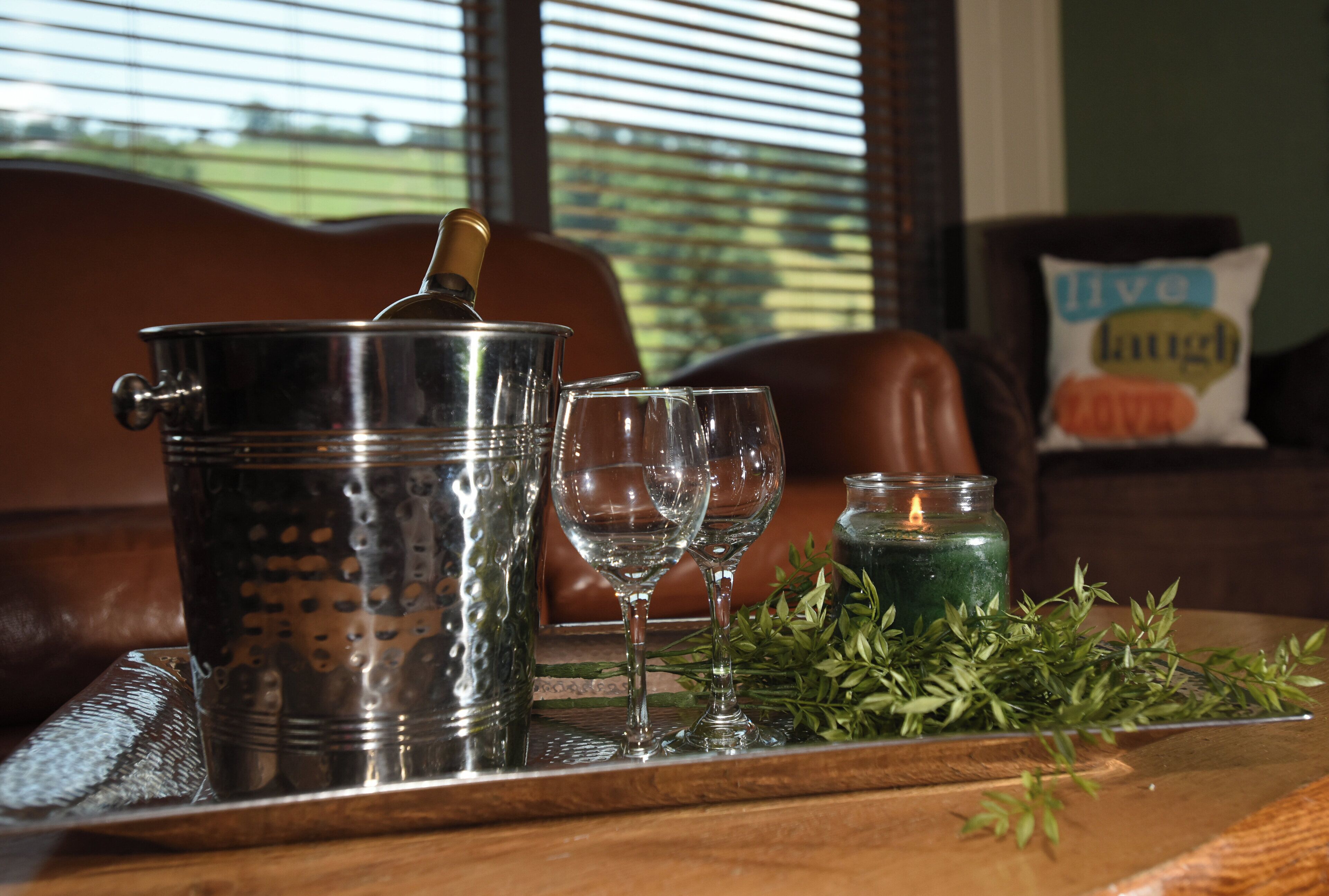 A metal container with a lid, two empty glasses, a lit candle, and green foliage on a wooden table.