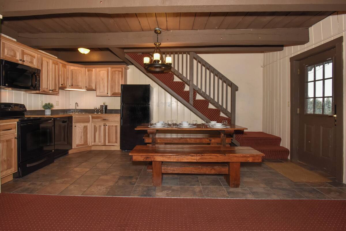 A kitchen with wooden cabinets, a black refrigerator, a wooden dining table set for four, and a staircase leading to a landing.