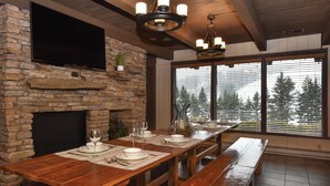 A dining area with a stone fireplace, wooden table, benches, and a large window with a view of trees. - Seven Springs Resort  by Vail Resorts (Champion)