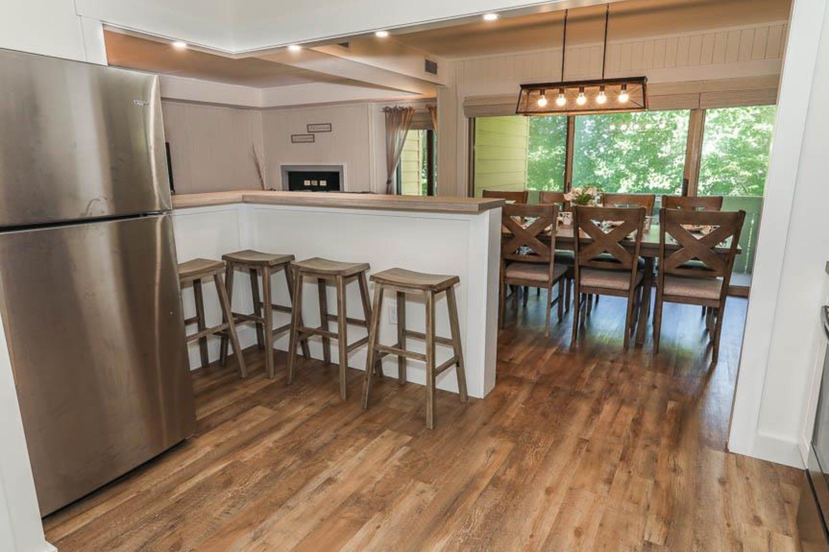 A kitchen with a stainless steel refrigerator, wooden stools, and a dining area with wooden chairs.