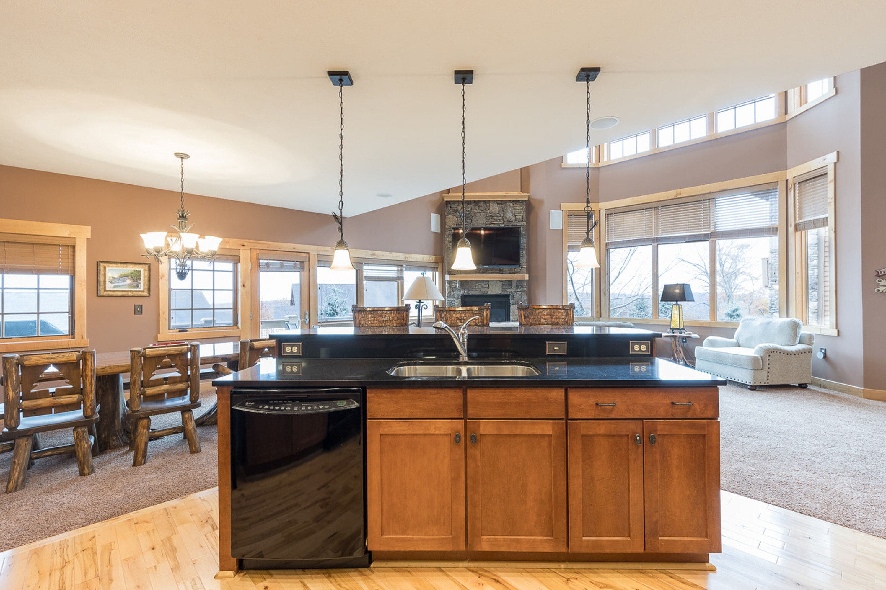 A spacious kitchen with a large island, wooden cabinets, and a built-in oven. There are hanging lights and a view of the outdoors through large windows.