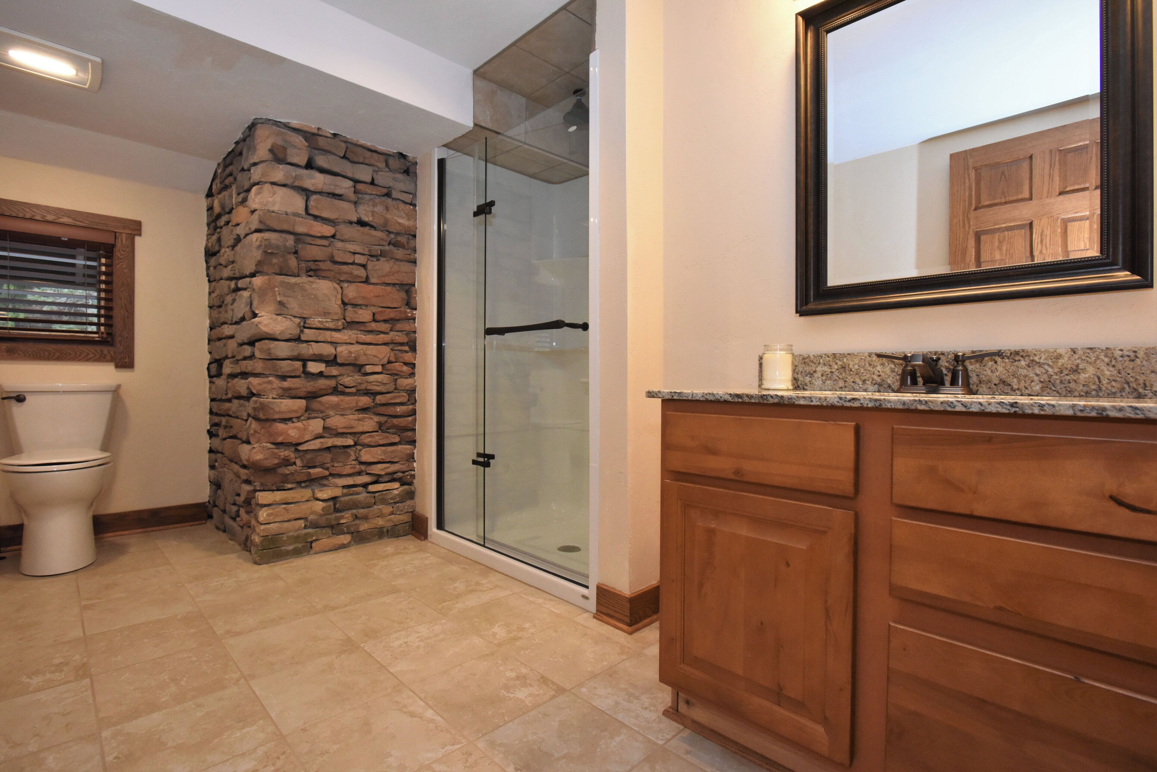 A bathroom with a stone accent wall, a glass shower enclosure, a toilet, wooden cabinets, and a mirror.