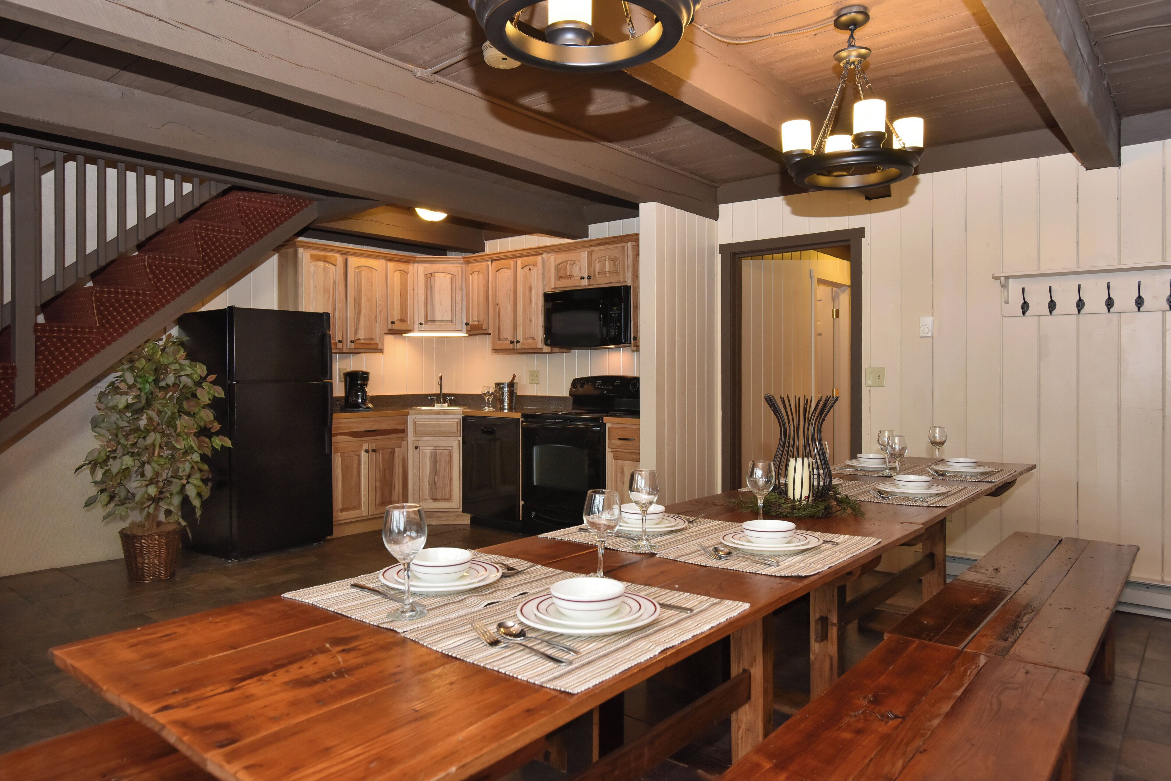 A wooden dining table set with plates, glasses, and cutlery, with a staircase and kitchen in the background.