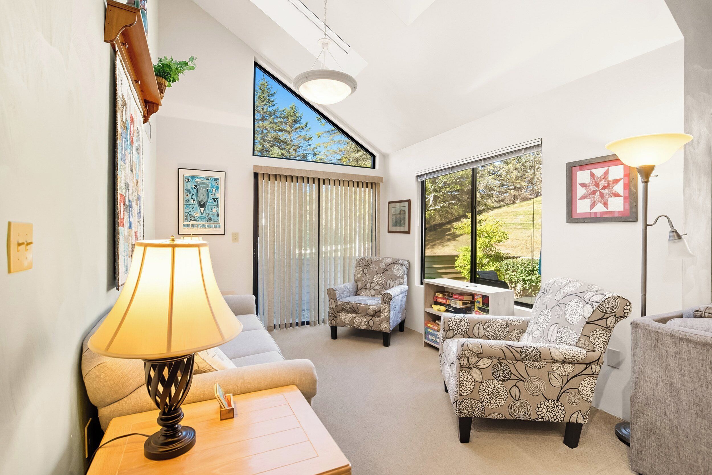 A spacious living room with a sofa, armchair, and a table with a lamp. There are large windows with blinds, a ceiling light, and a plant on the wall.
