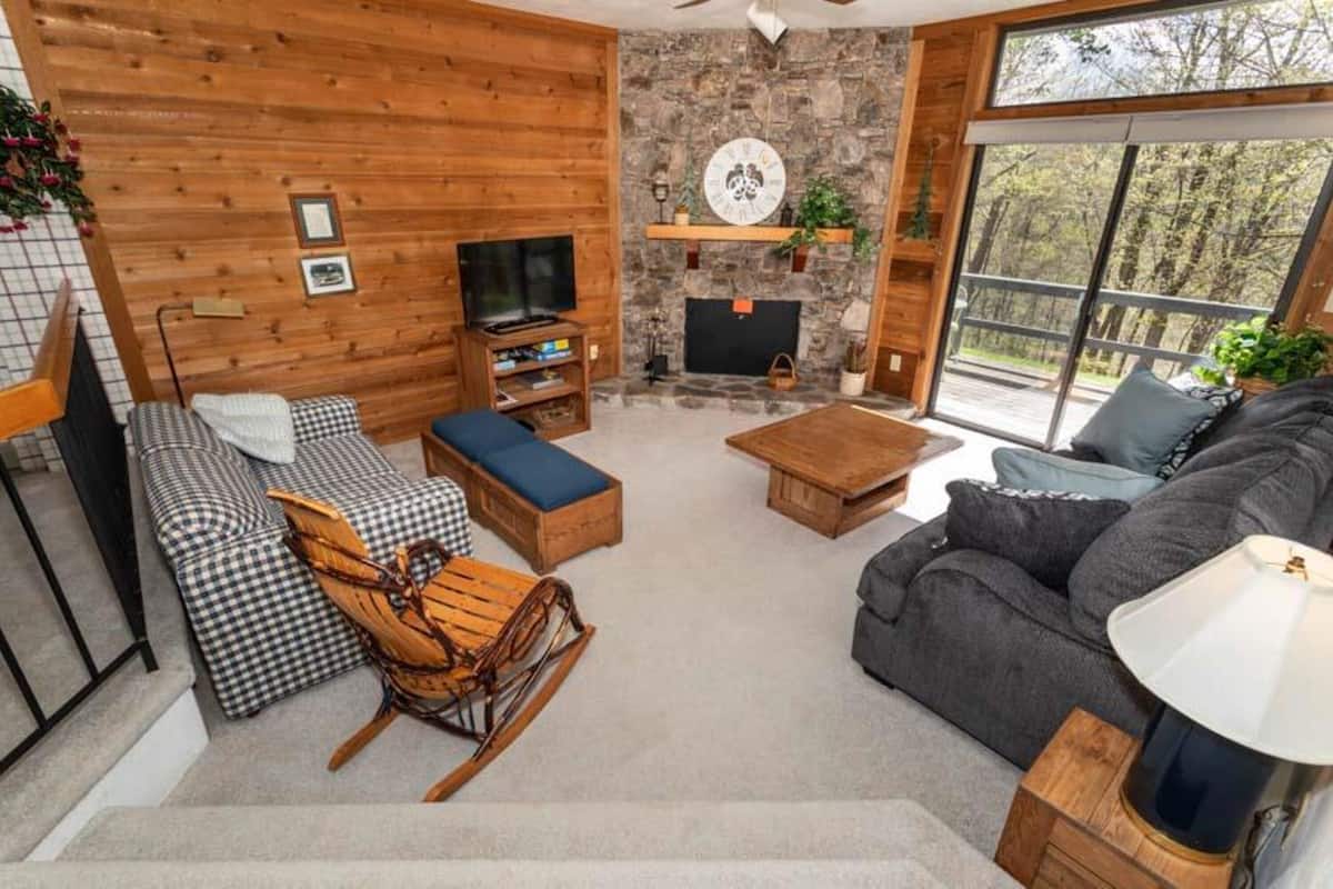 A cozy living room with a stone fireplace, wooden walls, and a large window overlooking a forest.