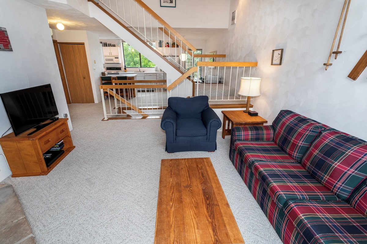 A living room with a sofa, a chair, a coffee table, a TV, and a staircase leading to a kitchen.