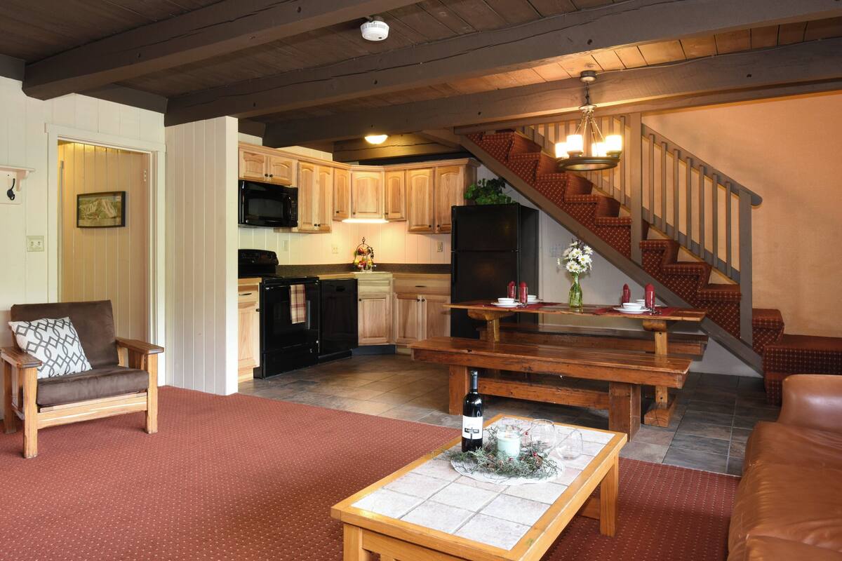 A spacious living area with a wooden bench, a coffee table, and a dining table. A staircase with a wooden railing leads to an upper level.