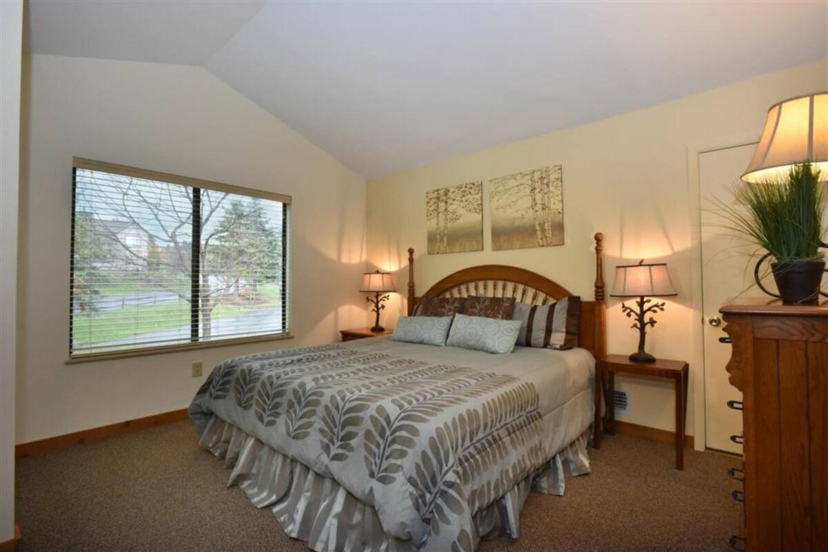 A bedroom with a large bed, two bedside lamps, a nightstand, a window with blinds, and a potted plant.