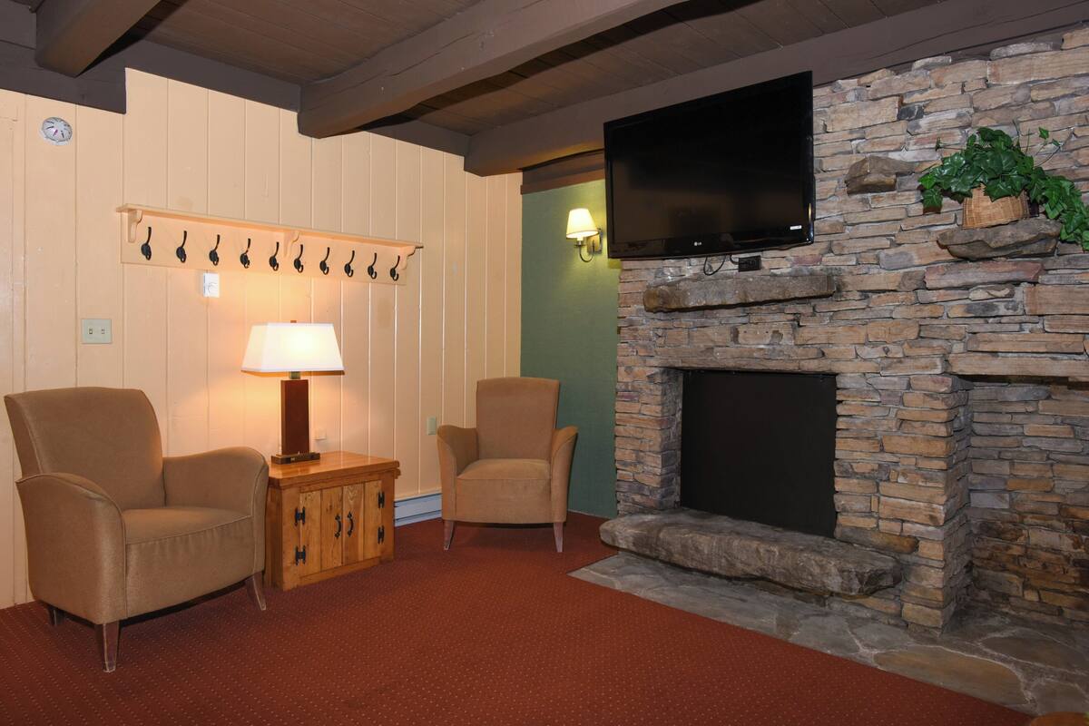 A cozy living room with two armchairs, a small wooden side table with a lamp, a stone fireplace, and a mounted flat-screen TV.