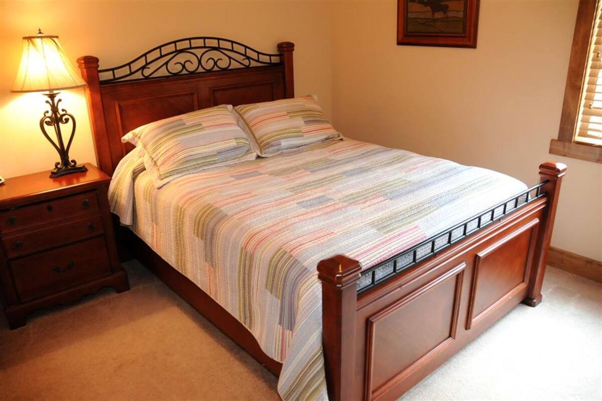 A neatly made bed with a striped bedspread, a wooden headboard, a nightstand with a lamp, and a small drawer.
