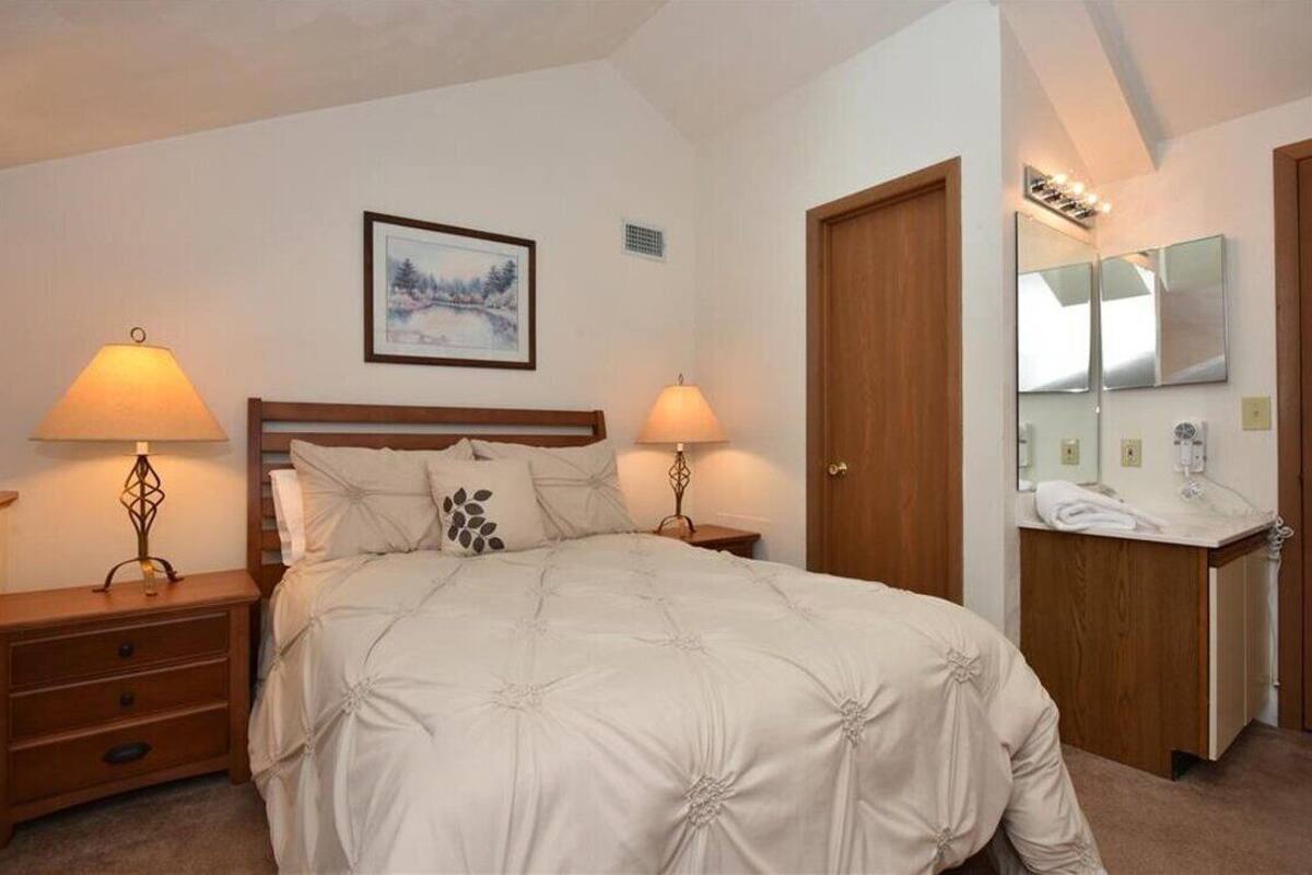 A neatly arranged bedroom with a large bed, two bedside lamps, a wooden nightstand, a framed picture on the wall, and a bathroom with a mirror and sink.
