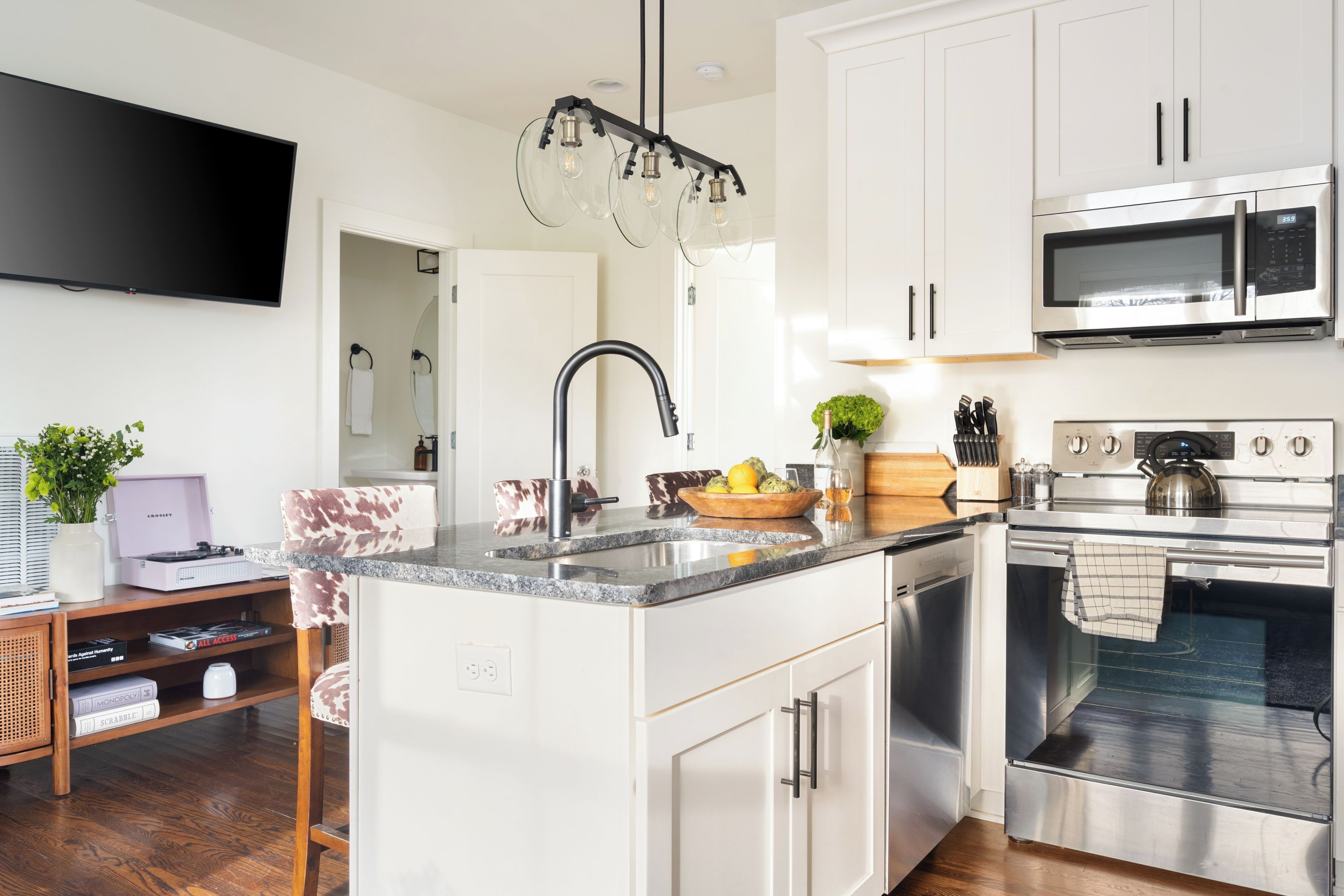 A modern kitchen with white cabinets, a stainless steel microwave, an oven, a granite countertop, and a built-in TV.