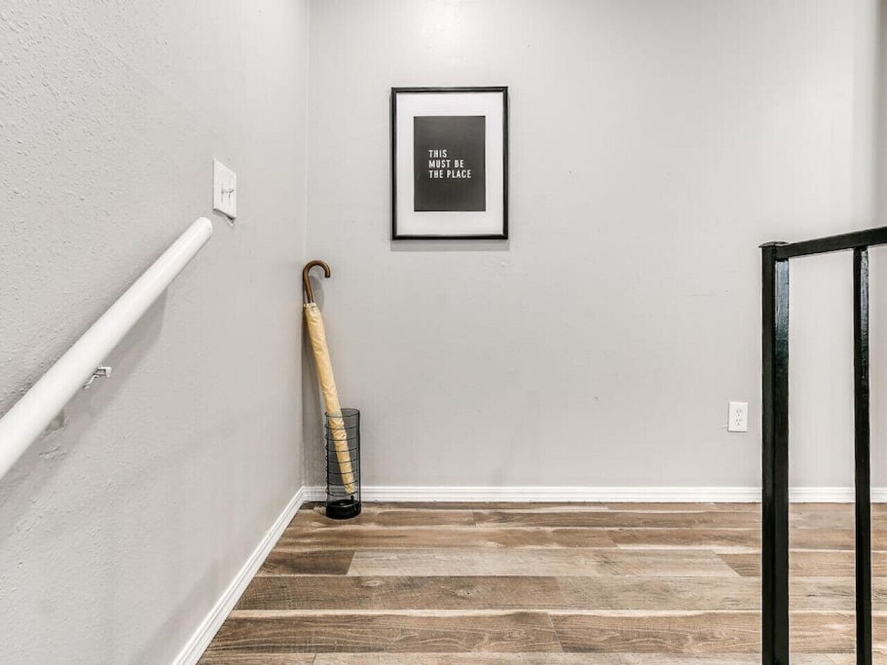 A hallway with a wooden floor, a white wall, a framed quote, and a cane leaning against the wall.